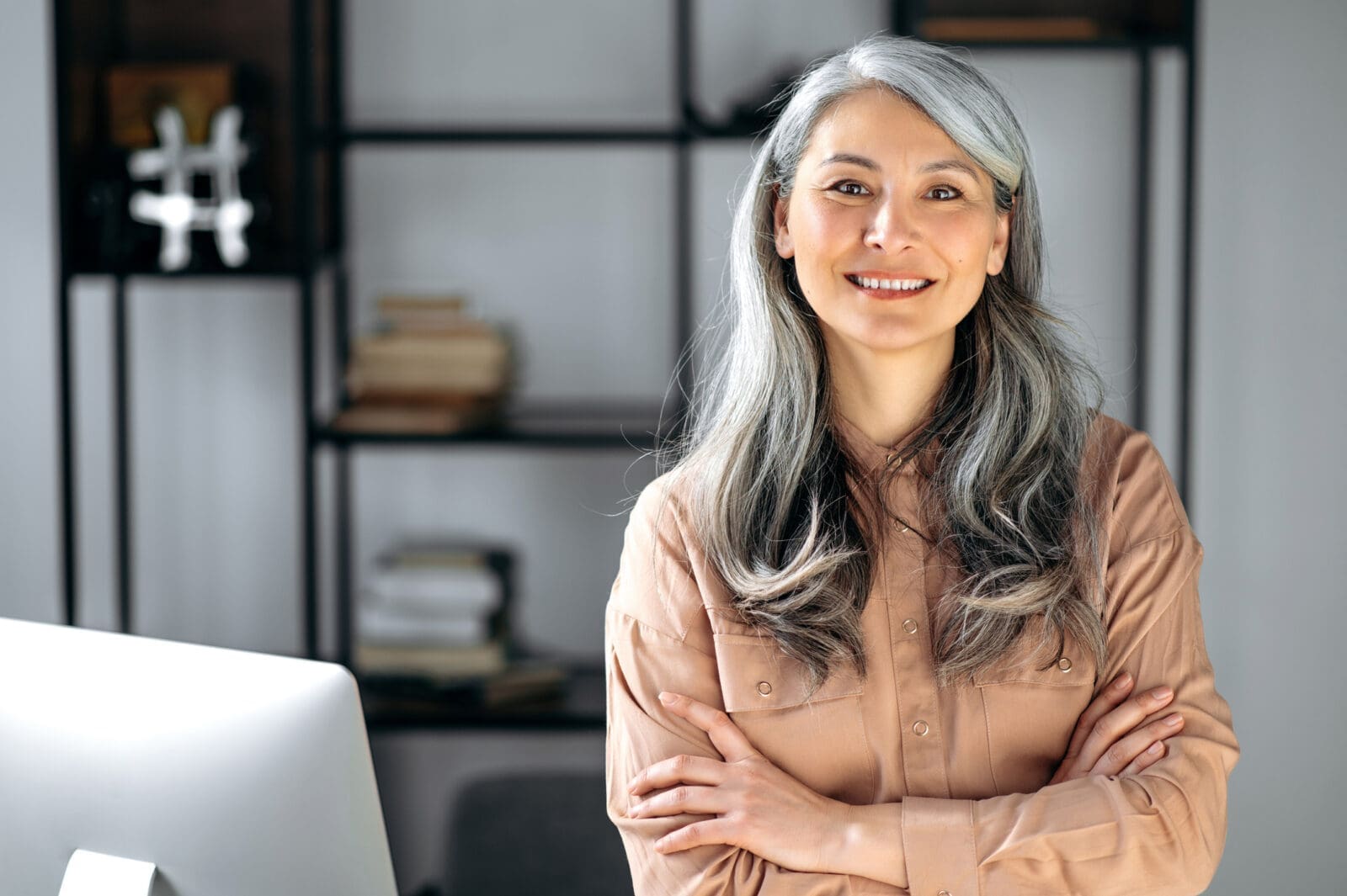 A smiling grey-haired Asian woman standing in her office beside a computer, smiling. CREDIT: https://www.istockphoto.com/portfolio/KaterynaOnyshchuk